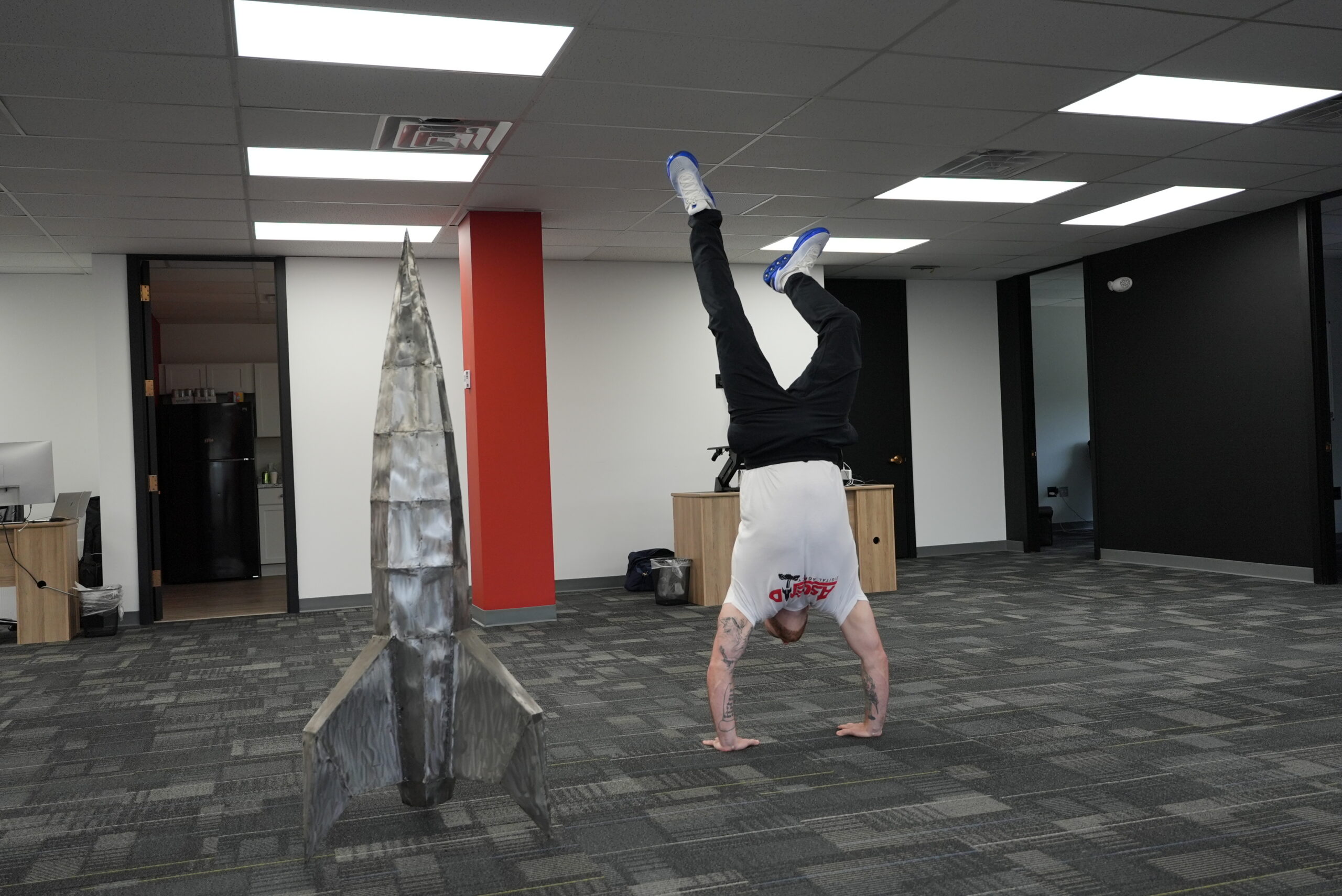 Person performing a handstand near a metallic rocket sculpture indoors.