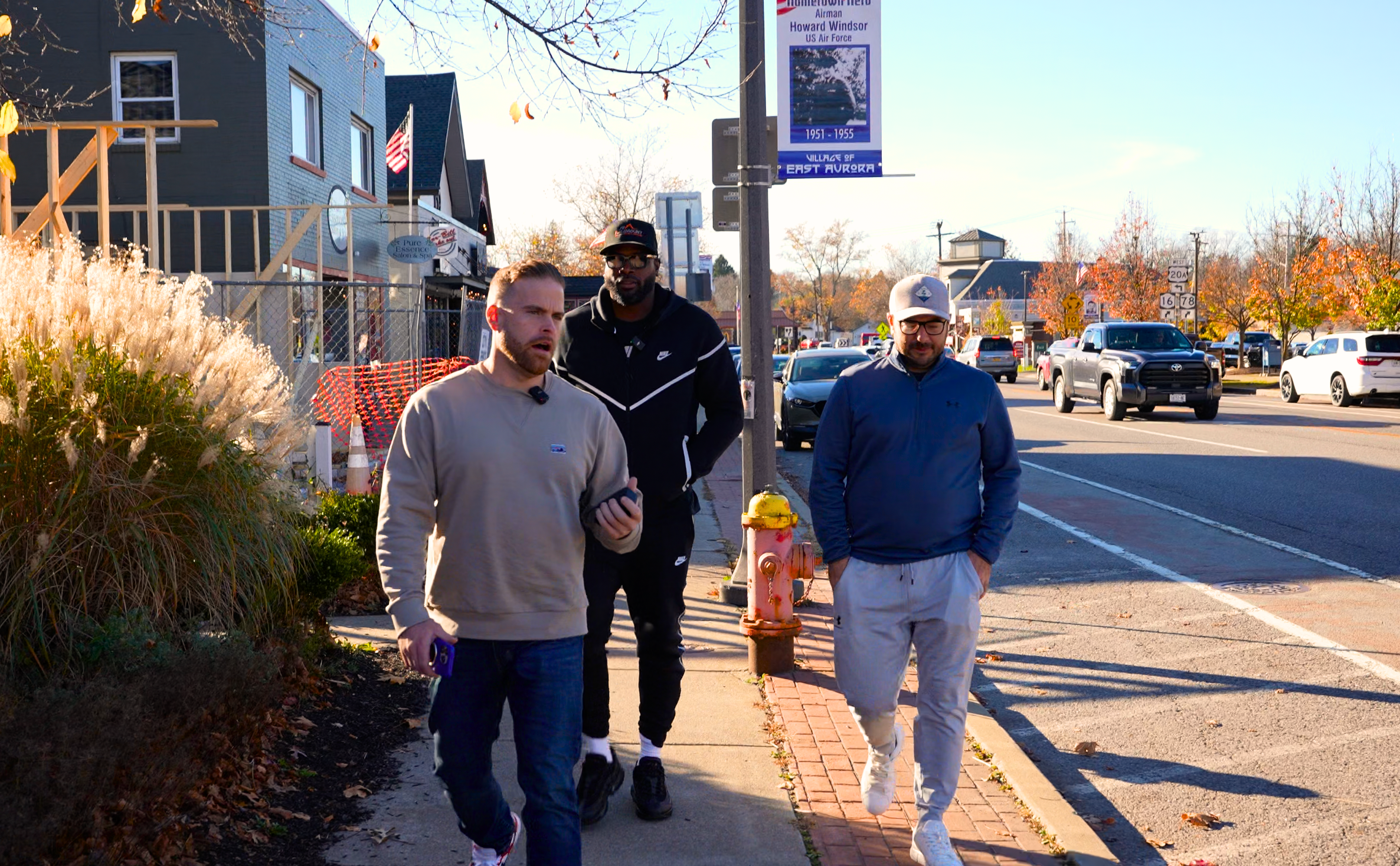 Three men walking on a sunny sidewalk in a suburban neighborhood with fall foliage.
